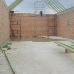Budowa domu murowanego: widok wnętrza z częściowo wzniesioną więźbą dachową, rusztowaniami i pracownikiem na górze, ściany z cegły, surowy beton na podłodze, drewniane elementy konstrukcyjne...