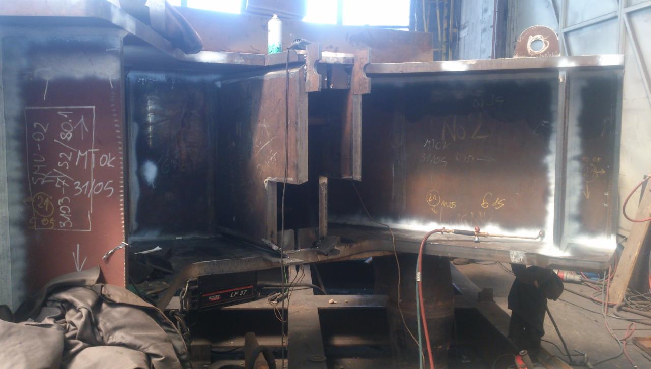 Konstrukcja stalowa w trakcie obróbki, widoczne oznaczenia kredą i spawarka Lincoln LF-37, surowe wykończenie metalu.
