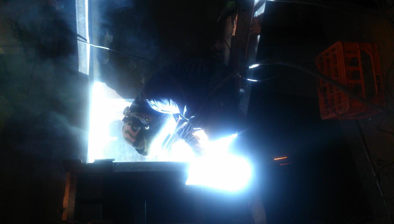 Praca w ciemnym otoczeniu, spawanie metalowej konstrukcji z intensywnym błyskiem światła, w tle widoczna pomarańczowa skrzynka.