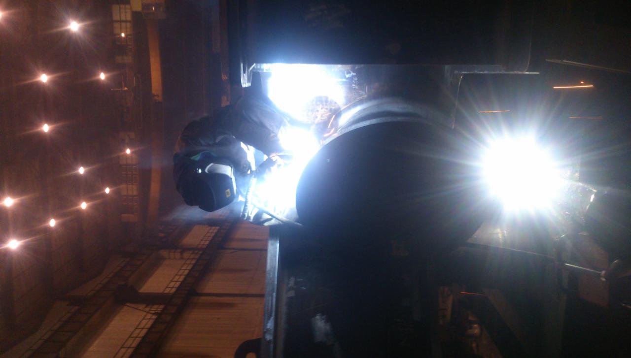 Praca przy spoinie rurociągu w hali przemysłowej, iskry i intensywne światło podczas procesu łączenia metalu.