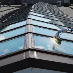 Czyszczenie szklanego dachu budynku za pomocą szczotki na długim, niebieskim wysięgniku; widoczne krople wody i odbicie nieba w czyszczonych szybach.