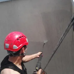 Pracownik w czerwonym kasku, zabezpieczony linami, czyści elewację budynku za pomocą myjki ciśnieniowej z bliska, widoczne ślady spływającej wody.