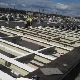 Pracownik w kamizelce odblaskowej maluje metalową konstrukcję dachu na budowie z widokiem na miasto w tle.