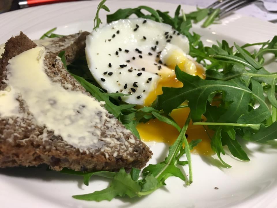 Talerz z połówką jajka w koszulce posypanego czarnuszką, kromką chleba żytniego posmarowaną masłem i świeżą rukolą.