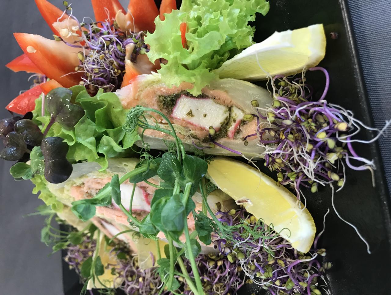 Delikatne roladki z łososia, kraba i pesto, udekorowane świeżymi sałatami, kiełkami i cząstkami cytryny, podane na czarnym talerzu.