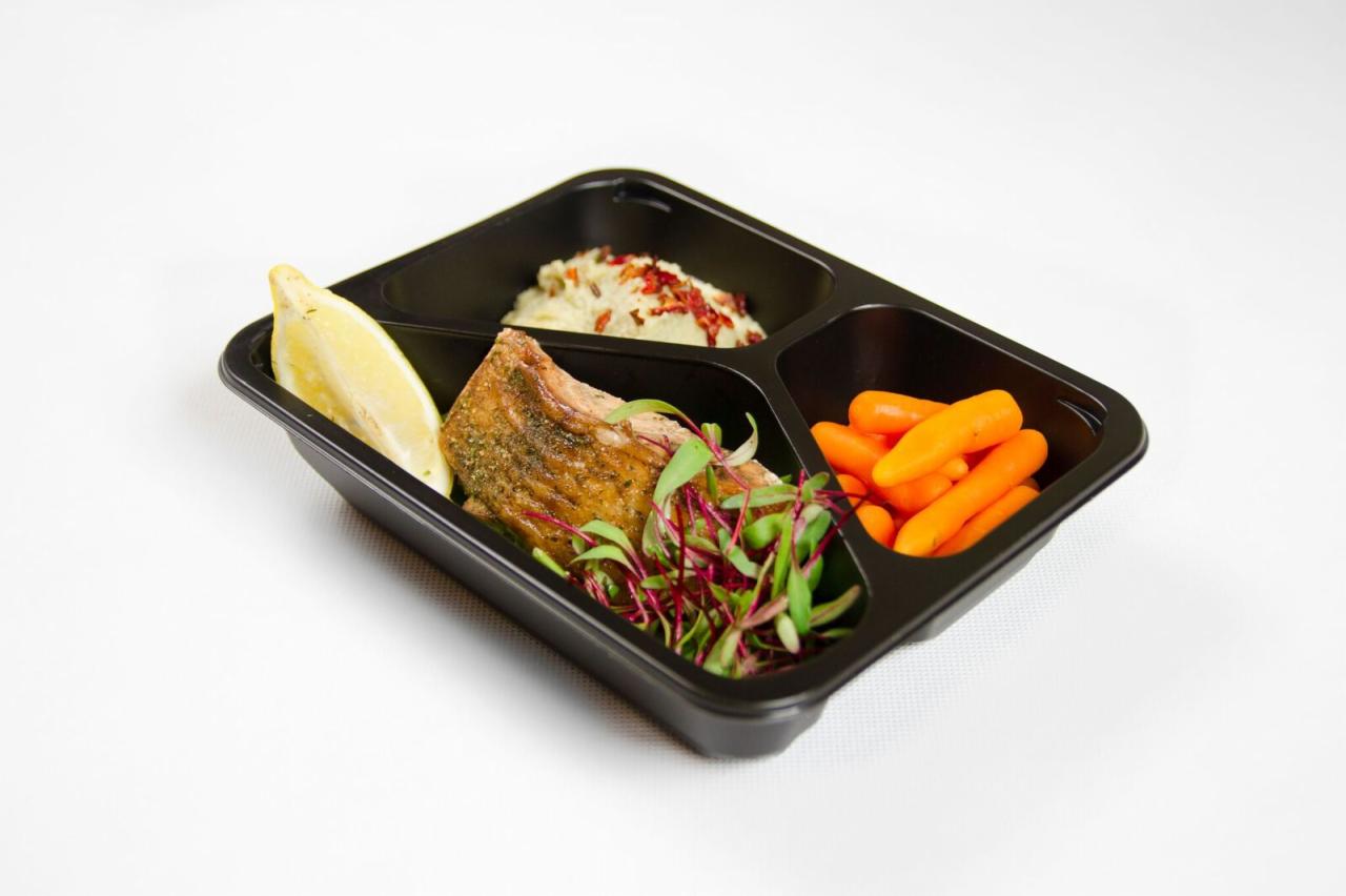 Czarna tacka z trzema przegródkami zawierająca porcję ryby z cytryną i mikroliśćmi, puree ziemniaczanym z czerwoną posypką oraz marchewką baby na białym tle.