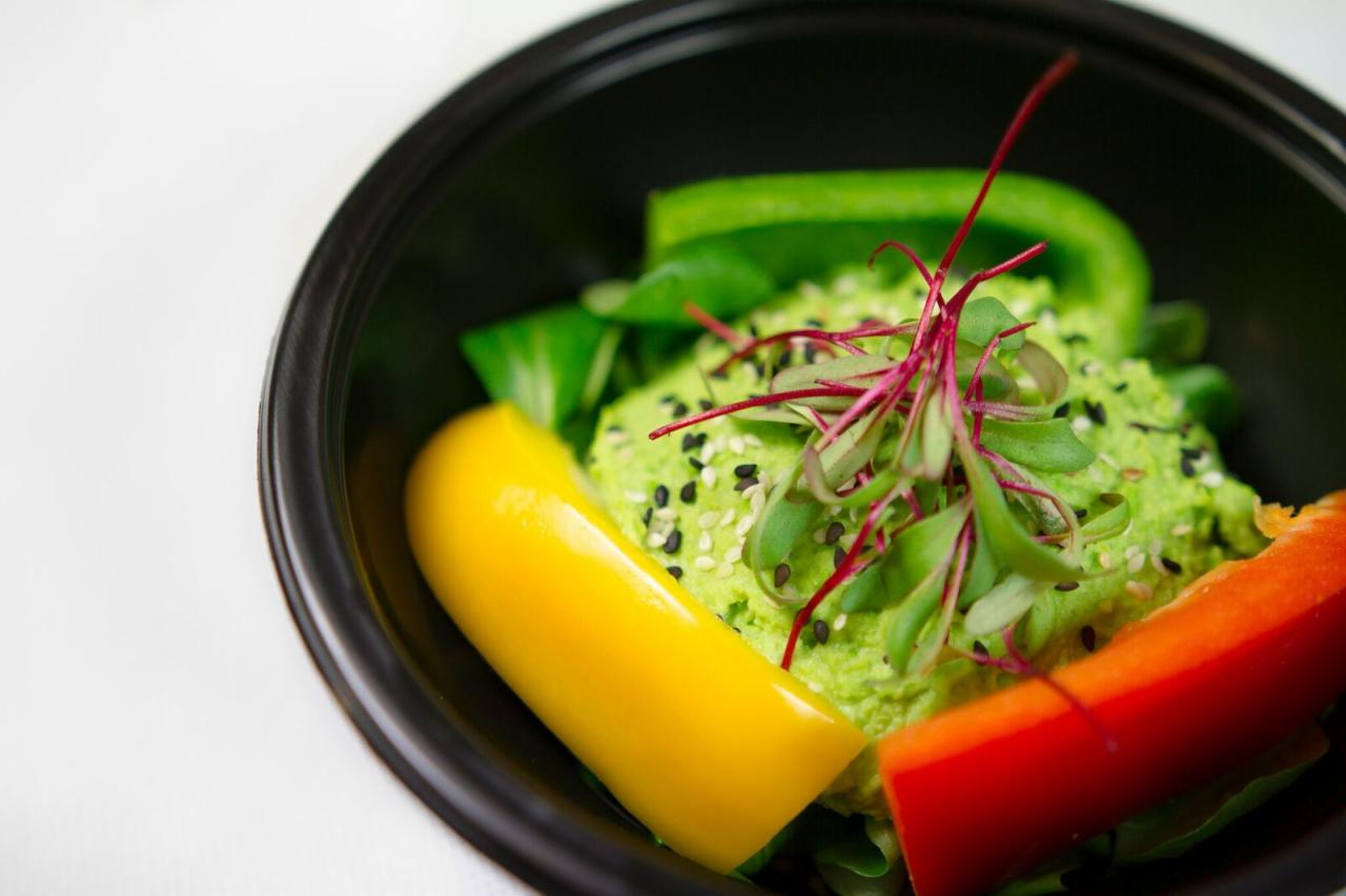 Porcja zielonej pasty z nasionami sezamu i mikro-ziołami, otoczona kawałkami kolorowej papryki w czarnym, okrągłym pojemniku na jasnym tle.