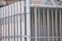 Fragment srebrnego ogrodzenia z metalowych profili pionowych i poziomych, połączonych klamrami, z widocznym słupkiem mocującym.