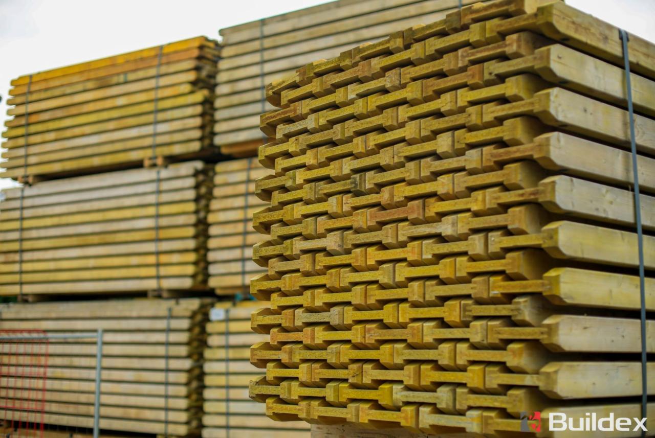 Stosy impregnowanych, drewnianych słupków ogrodzeniowych, ułożonych w równe warstwy i zabezpieczonych taśmą, w tle widoczne kolejne palety z materiałem.