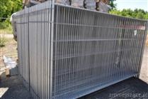 Stos ocynkowanych, spawanych paneli ogrodzeniowych, widoczne złącza i niewielka etykieta na jednym z paneli, na tle drzew i sterty drewna.