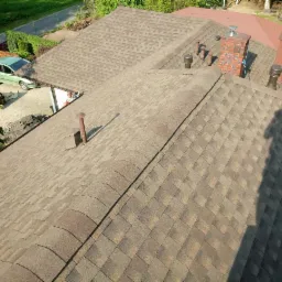 Widok z góry na brązowy dach pokryty gontem bitumicznym z kominem z czerwonej cegły i wentylacją, na tle zieleni i samochodu