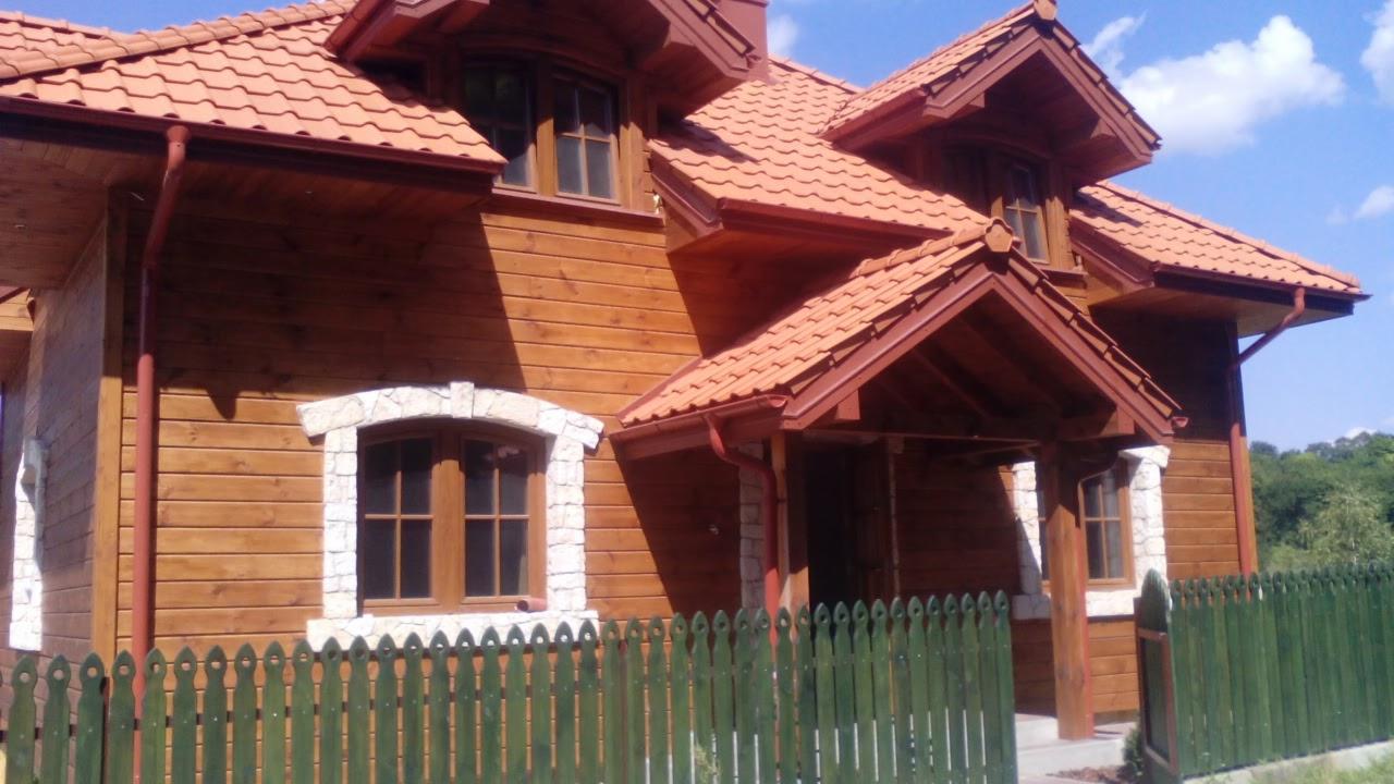 Drewniany dom z czerwoną dachówką, widoczne okna z kamienną obudową i zielone ogrodzenie.