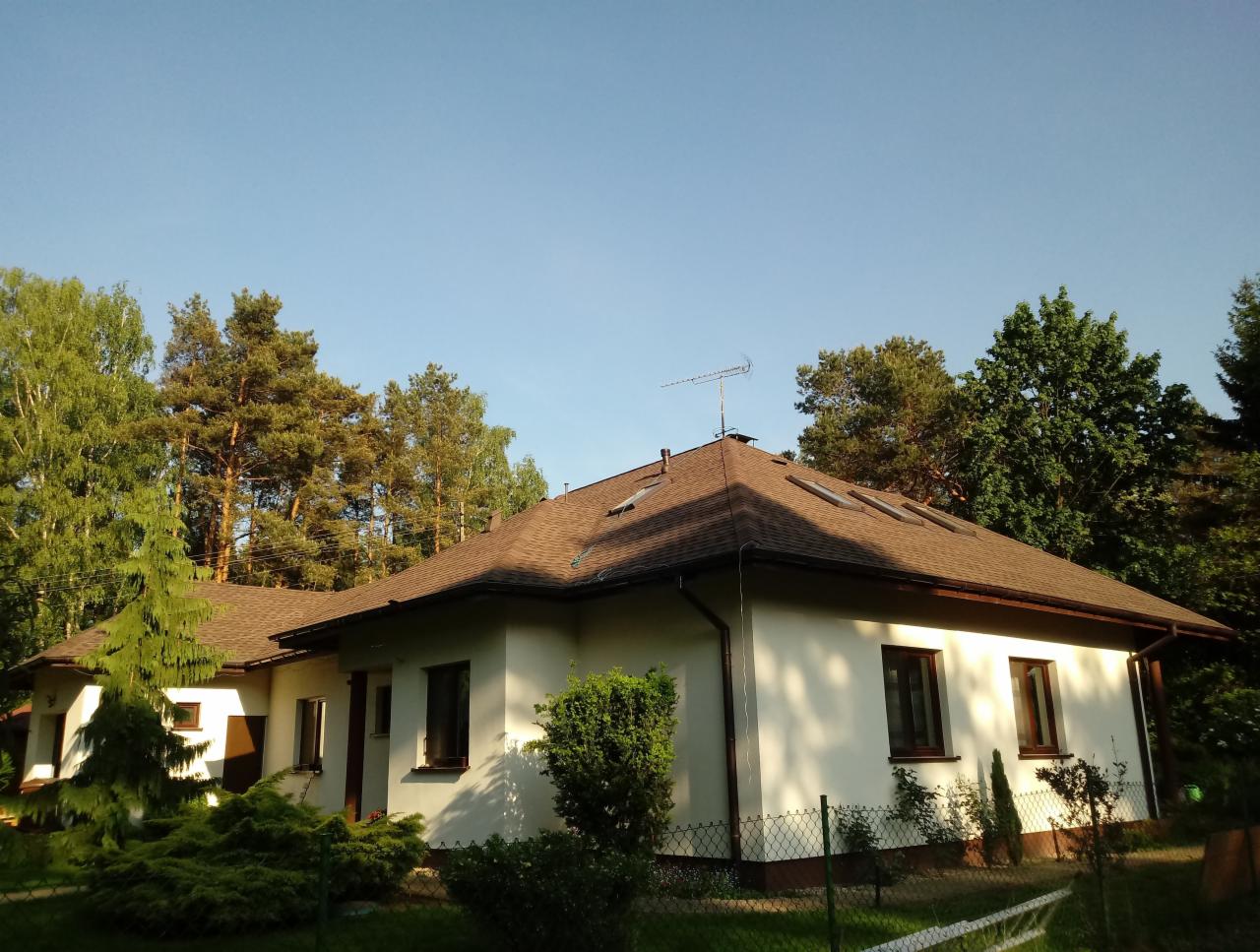 Dom jednorodzinny z brązowym dachem wielospadowym i oknami dachowymi, otoczony zielenią i drzewami iglastymi.