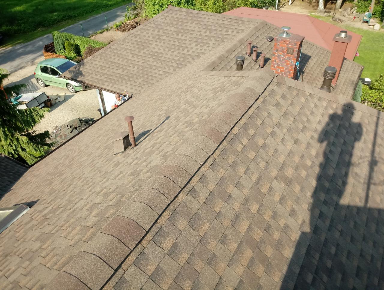 Widok z góry na brązowy dach pokryty gontem bitumicznym z kominem z czerwonej cegły i wentylacją, na tle zieleni i samochodu
