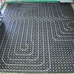 Rozłożona instalacja ogrzewania podłogowego z białych rurek na czarnej macie z otworami, widoczne niebieskie elementy mocujące instalację.