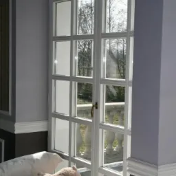 Białe, przeszklone drzwi balkonowe z widokiem na balkon z kamienną balustradą i drzewa, wnętrze pokoju z pluszowymi zabawkami na kanapie.