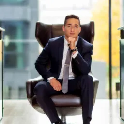 Młody mężczyzna w granatowym garniturze i szarym krawacie siedzi w skórzanym fotelu na tle nowoczesnego biura z dużymi oknami.