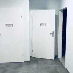 Dwa białe skrzydła w korytarzu: jedno zamknięte z napisem 'Pomieszczenie gospodarcze', drugie uchylone z napisem 'Toaleta dla personelu', szare płytki na podłodze.