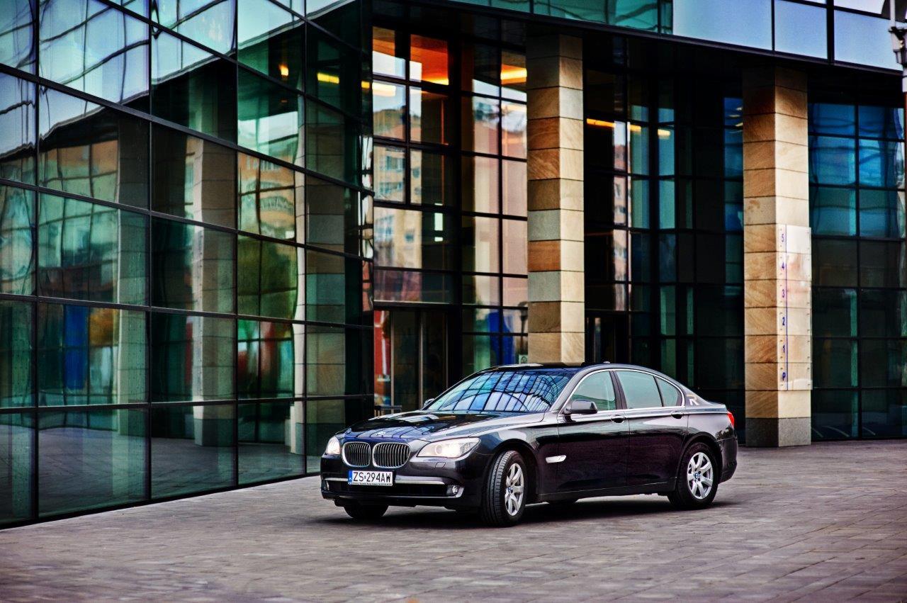 Elegancka czarna limuzyna BMW serii 7 zaparkowana przed nowoczesnym biurowcem ze szklaną fasadą, widoczne odbicia w szybach budynku.