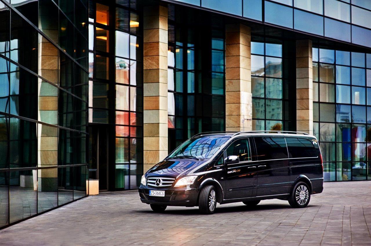 Czarny van Mercedes-Benz Viano z przyciemnianymi szybami zaparkowany przed nowoczesnym budynkiem biurowym z szklaną fasadą i kamiennymi kolumnami.