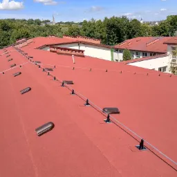 Czerwony dach pokryty papą termozgrzewalną z systemem odgromowym, na tle zieleni i zabudowań. Widok z góry, słoneczny dzień.