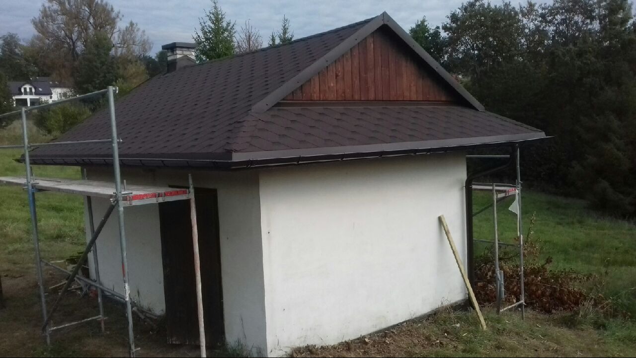 Mały budynek z brązowym dachem bitumicznym i drewnianym szczytem, widoczne rusztowanie z lewej strony, tło stanowią drzewa i budynek w oddali.