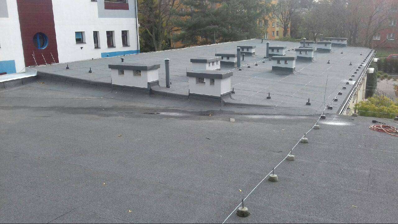 Widok z góry na płaski, pokryty papą dach budynku z licznymi wywietrznikami, zabezpieczony odgromowo. W tle zabudowa miejska i drzewa.