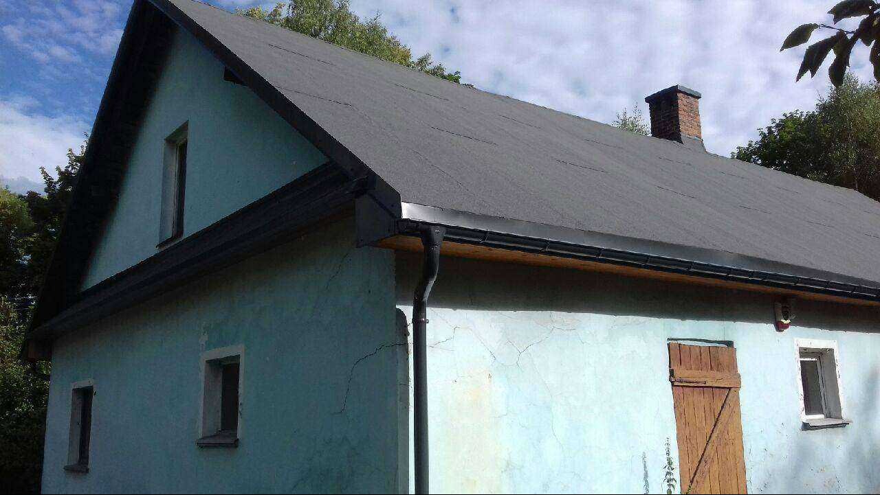 Dom z nowym, ciemnoszarym dachem z papy bitumicznej i ciemnymi rynnami. Widoczny komin z cegły. Ściany budynku w odcieniu błękitu z widocznymi spękaniami.