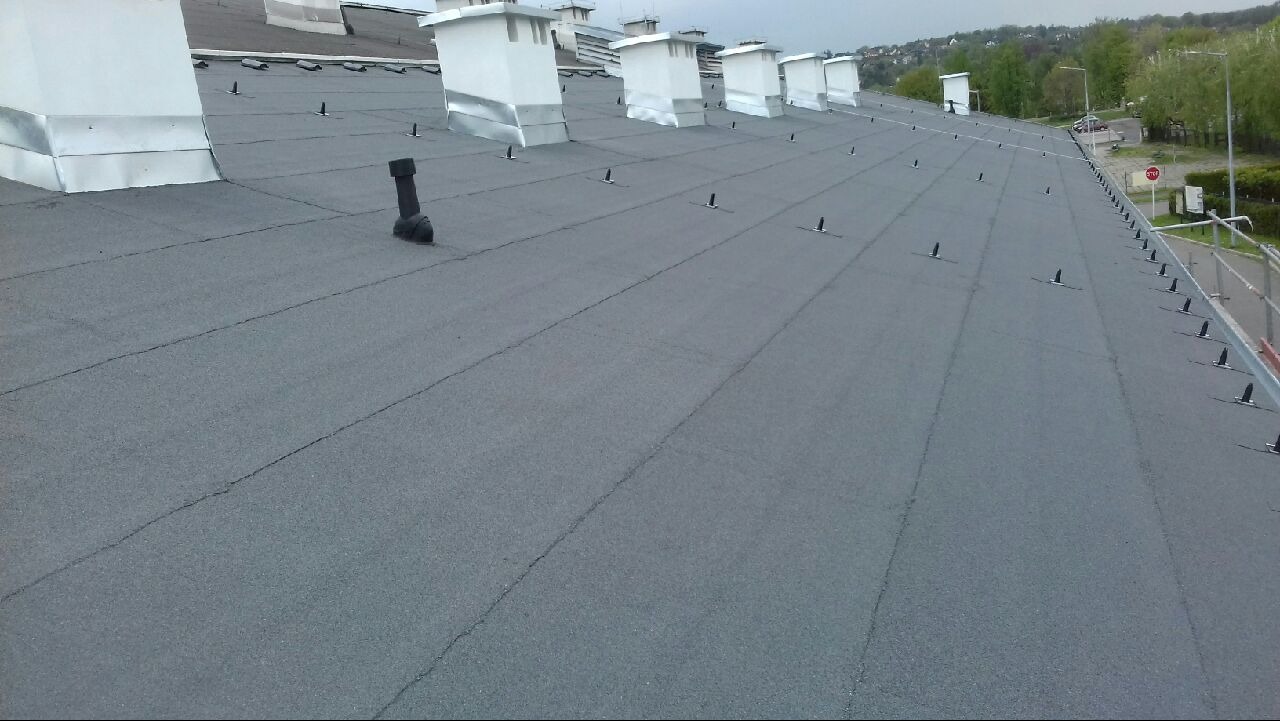 Szary dach pokryty papą termozgrzewalną z kominami i mocowaniami, widok z góry ukazuje równą powierzchnię i detale wykończenia.