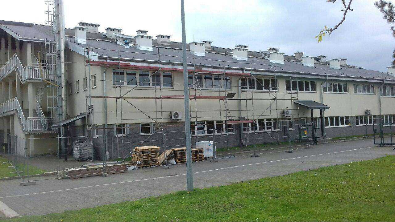 Budynek w trakcie remontu dachu; widoczne rusztowania, materiały budowlane i kominy. Szary dach z licznymi kominami.