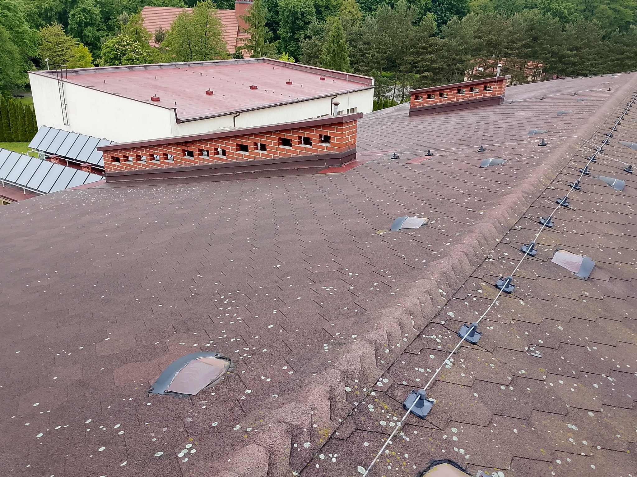 Dach pokryty brązowym gontem bitumicznym z widocznymi elementami systemu odgromowego i kominkami wentylacyjnymi. Widoczne zabrudzenia i porosty na powierzchni dachu.