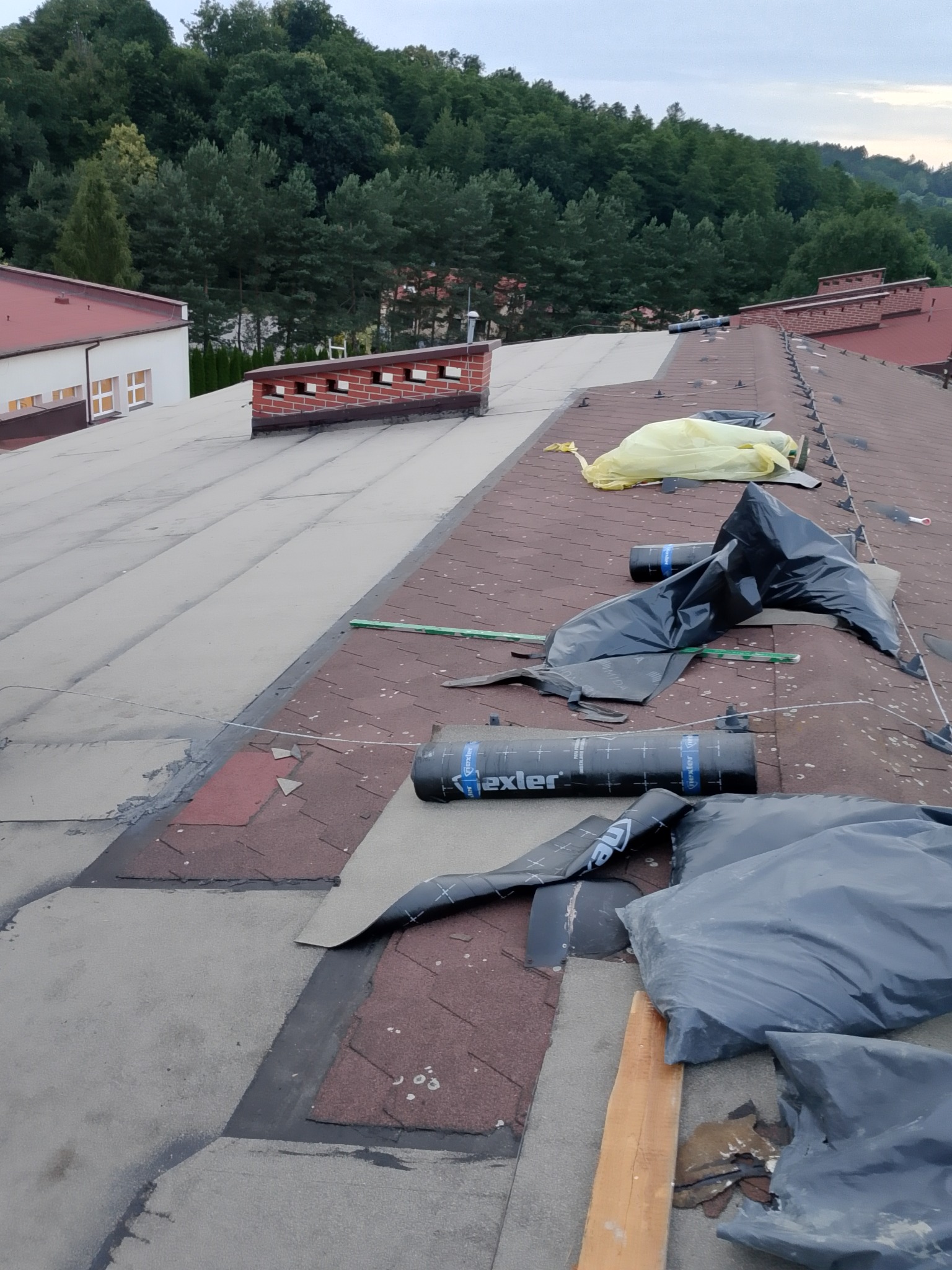 Dach pokryty papą, częściowo wykończony. Widoczne rolki papy, narzędzia i fragmenty starego pokrycia. W tle budynek i drzewa.