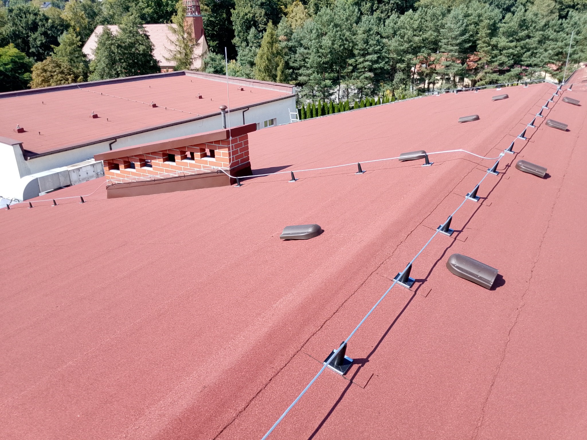 Czerwony dach z papy termozgrzewalnej z systemem odgromowym na budynku. Widoczny komin z cegły i elementy wentylacyjne. W tle drzewa.