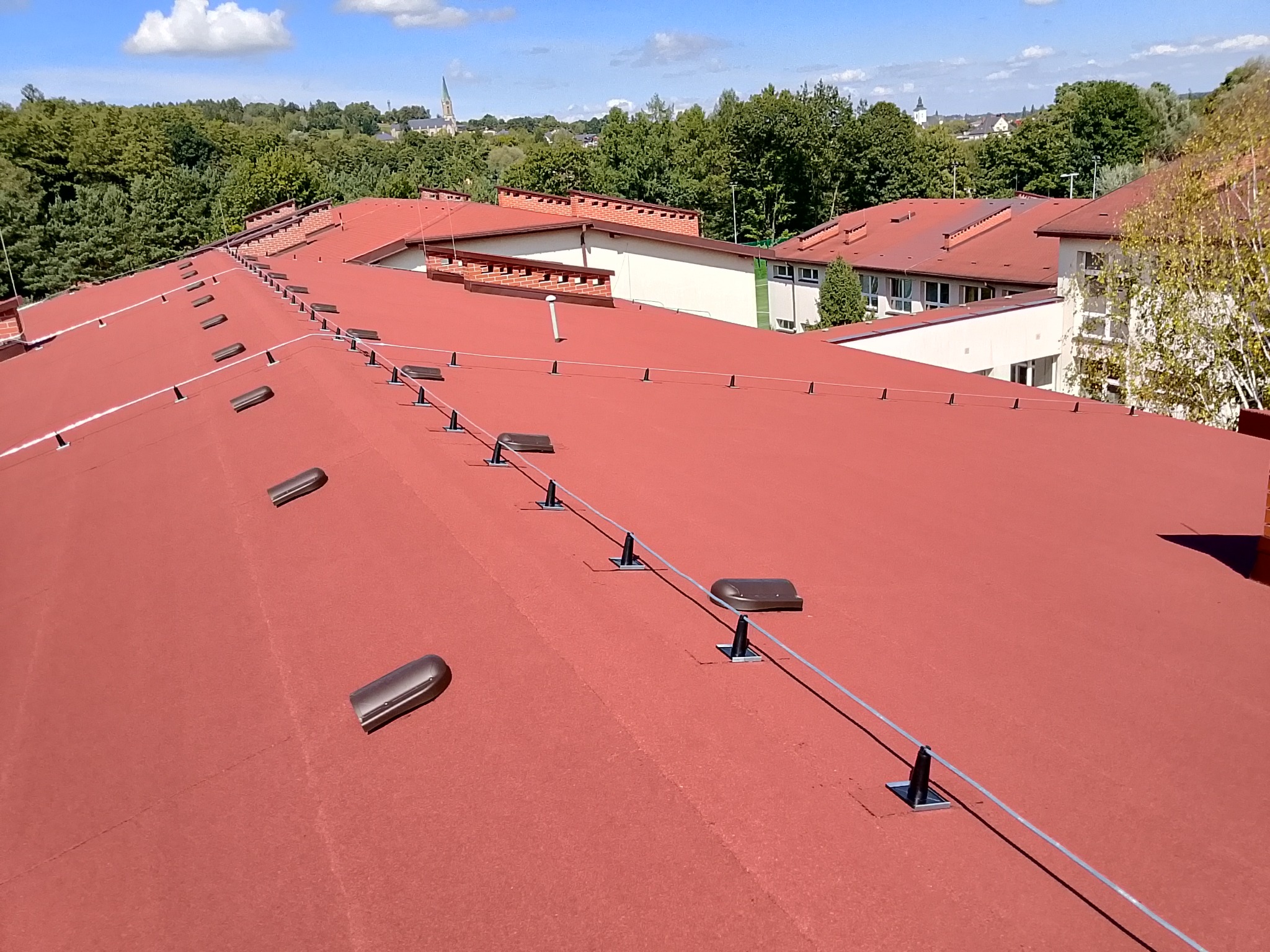 Czerwony dach pokryty papą termozgrzewalną z systemem odgromowym, na tle zieleni i zabudowań. Widok z góry, słoneczny dzień.