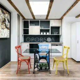 Jasna kuchnia z jadalnią w stylu rustykalnym, stół z blatem ze szkła na podstawie starej maszyny do szycia, krzesła w kolorach pomarańczowym, niebieskim i żółtym, drewniane belki na suficie, obraz...