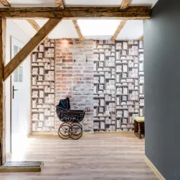 Wnętrze przedpokoju z drewnianą konstrukcją stropu i ścianą w cegły oraz tapetą z motywem książek, z antycznym wózkiem dziecięcym.