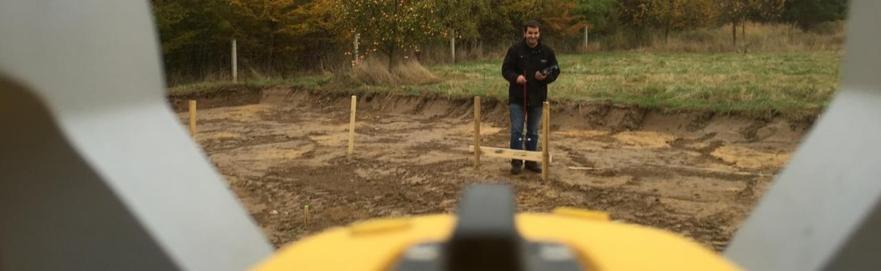 Pomiar geodezyjny działki pod budowę domu, widok z perspektywy instrumentu geodezyjnego, na drugim planie geodeta z tyczką.