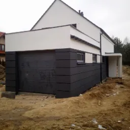 Nowoczesny dom z elewacją w kolorze grafitu i bieli, widok z zewnątrz, teren wokół w trakcie prac wykończeniowych.