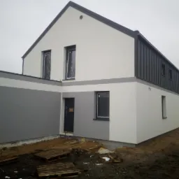 Dwukolorowa elewacja domu jednorodzinnego z szarymi akcentami, widoczne palety na ziemi, pochmurne niebo.