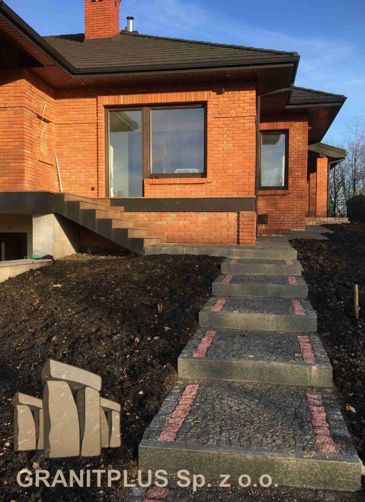 Schody wejściowe do domu wyłożone ciemną kostką z czerwonymi akcentami, prowadzące od trawnika do ceglanego budynku.