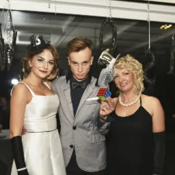 Trzy osoby ubrane na galowo: kobieta w białej sukience i czarnych rękawiczkach, mężczyzna w szarym garniturze z muchą i kobieta w czarnej sukience z perłami, trzymająca kostkę Rubika. Dekoracje...