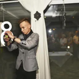 Młody iluzjonista w szarej marynarce i muszce, rozwiązuje kostkę Rubika na tle dekoracji urodzinowej osiemnastki i obserwujących go gości.