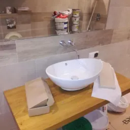 Biała umywalka nablatowa na drewnianym blacie w łazience w trakcie remontu, widoczne narzędzia i materiały budowlane, odbicie w lustrze z półkami i akcesoriami łazienkowymi.