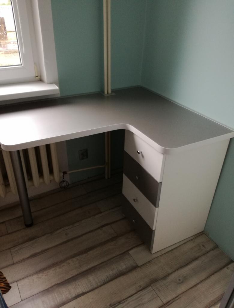 Narożne biurko z szufladami w pokoju, widok na grzejnik pod oknem i drewnopodobną podłogę.