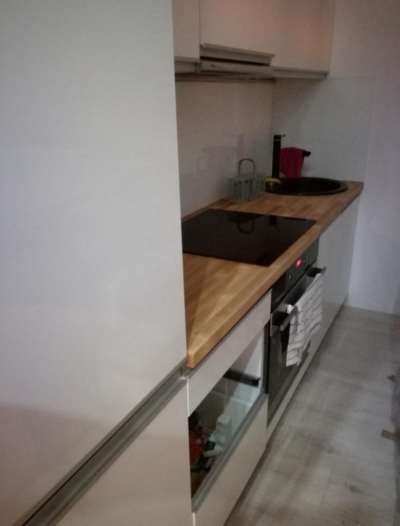 Niewielka kuchnia w minimalistycznym stylu z białymi frontami, drewnianym blatem, czarnym zlewem i płytą indukcyjną. Widoczny piekarnik z ręcznikiem na uchwycie.