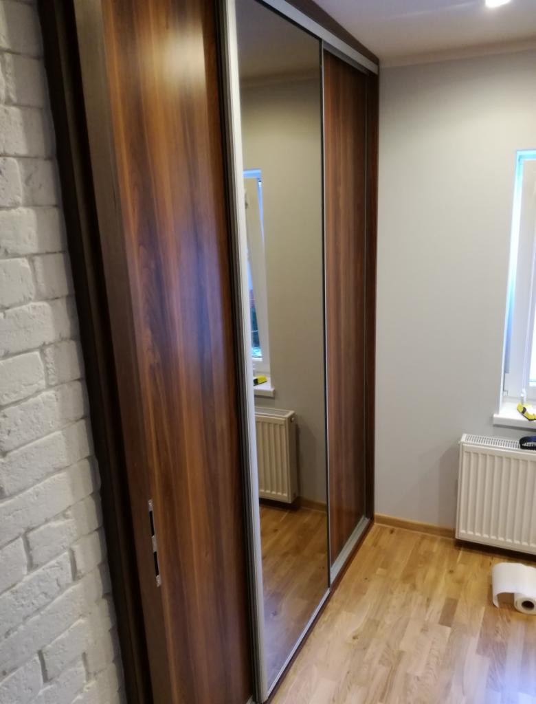 Wysoka szafa przesuwna z lustrem, drewnopodobne fronty, widoczna ściana z białej cegły i parkiet, w lustrze odbicie okna z grzejnikiem.