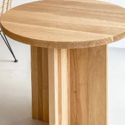 Okrągły stół z jasnego drewna z unikalną podstawą z pionowych paneli, obok krzesło z rattanu na metalowych nogach.