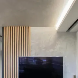 Nowoczesny salon z dekoracyjną ścianą z drewnianych listew i efektem stiuku w odcieniach szarości, oświetlenie punktowe i listwa LED na suficie.