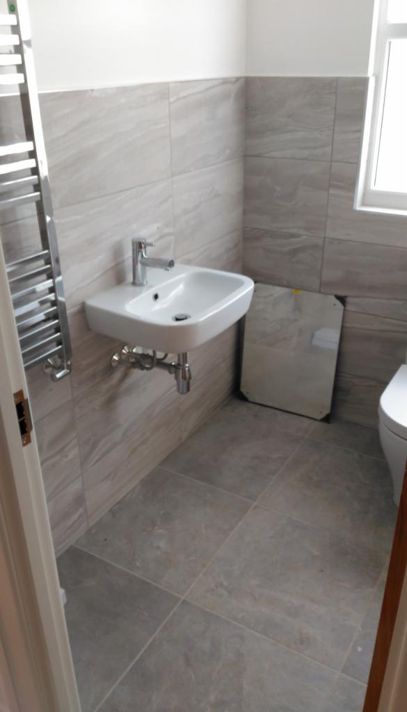 Wykończona łazienka z umywalką nablatową, grzejnikiem drabinkowym i dużymi, szarymi płytkami imitującymi kamień na ścianach i podłodze. Lustro leży oparte o ścianę przed montażem.