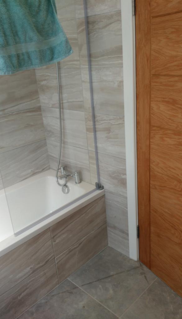 Wykończona łazienka z wanną obudowaną płytkami imitującymi drewno, szklanym parawanem nawannowym i turkusowym ręcznikiem.